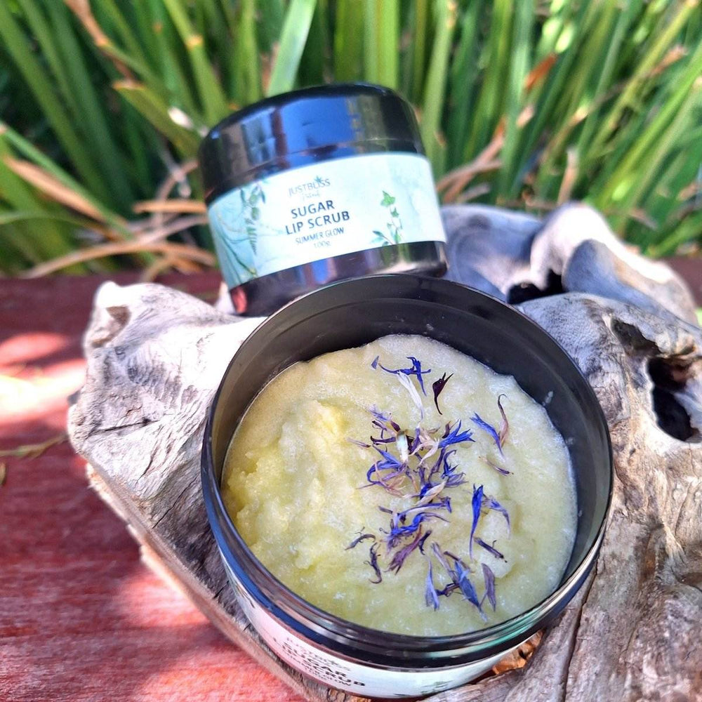 Summer Glow Sugar Lip Scrub in a jar, open to show yellow scrub with blue flower petals, on a rustic wooden surface.