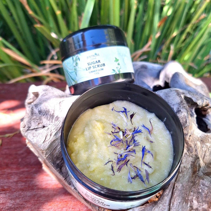 Summer Glow Sugar Lip Scrub in a jar, open to show yellow scrub with blue flower petals, on a rustic wooden surface.
